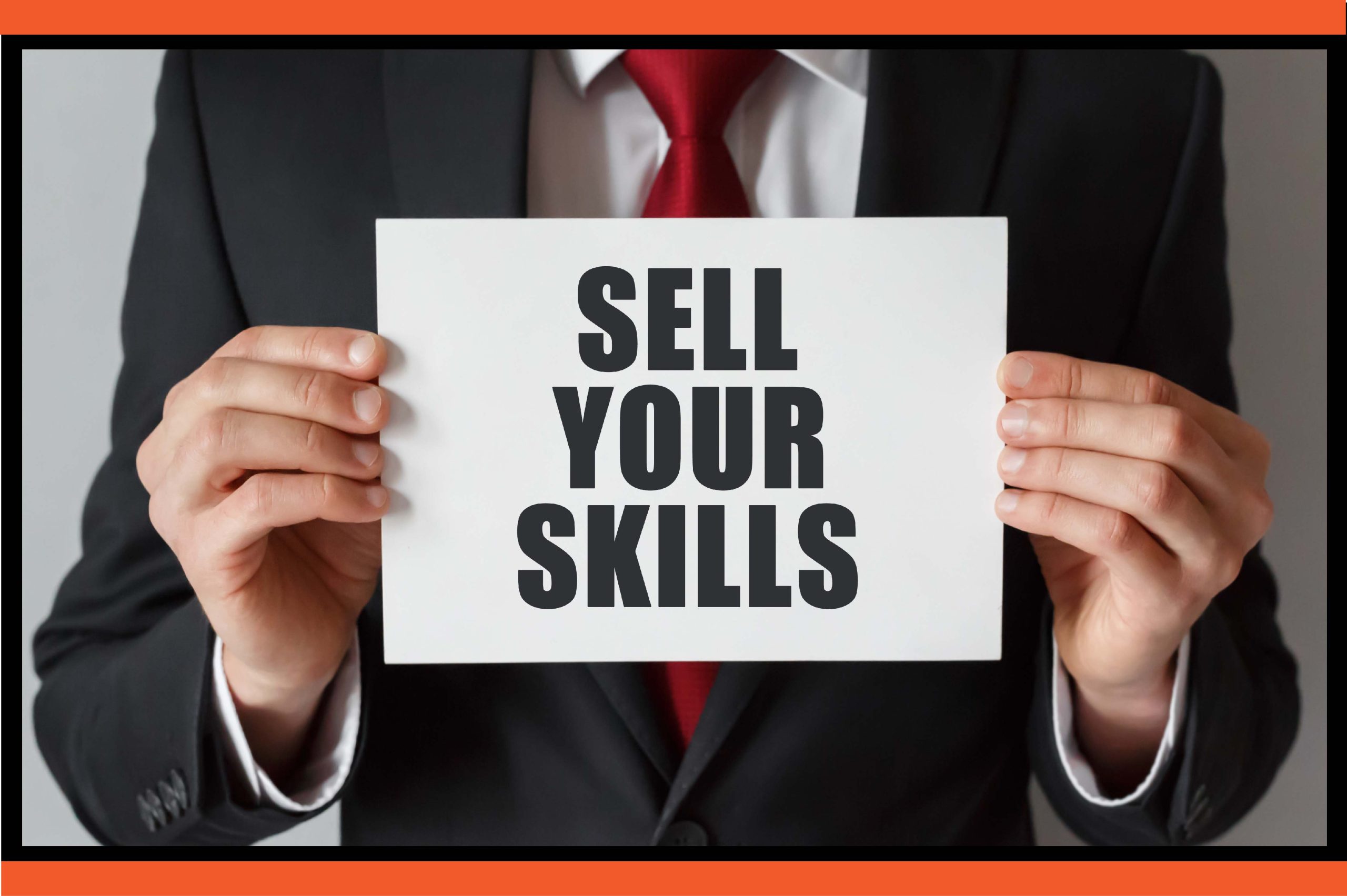 A headless picture of a guy in a dark suit with a white shirt and red tie holding an A4 white paper written SELL YOUR SKILLS